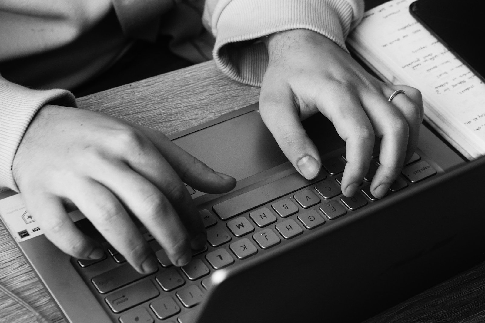 Black and white detail of hands working on a laptop beside handwritten notes