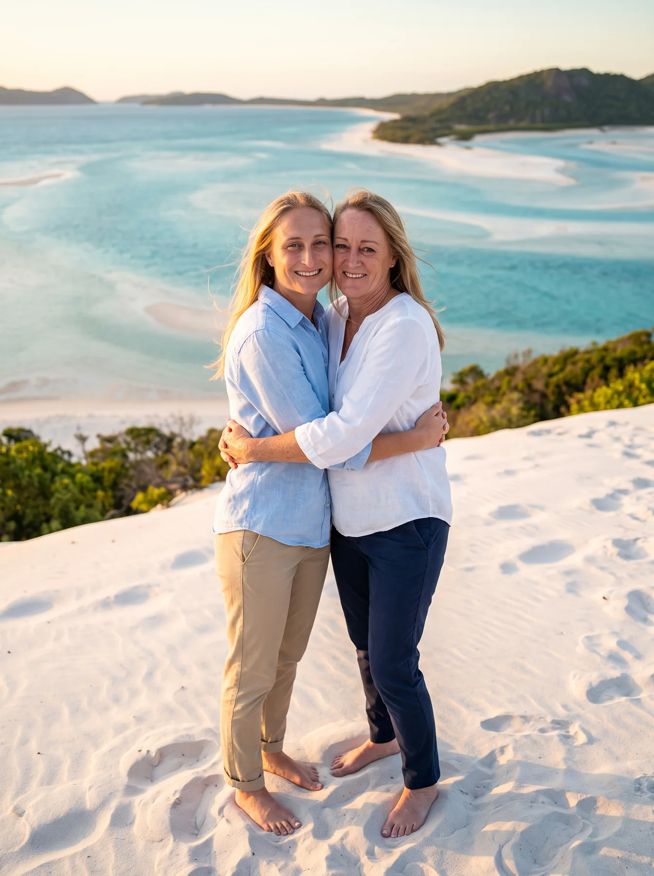 Two women embracing on white sand above turquoise water