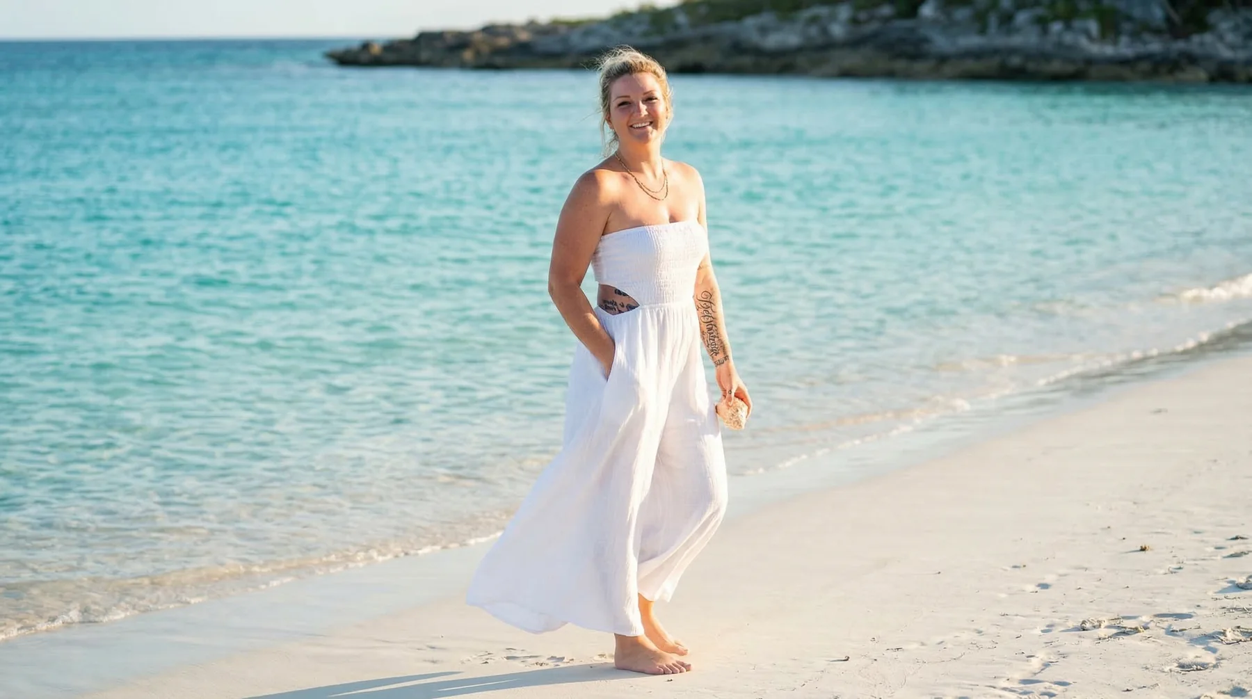 A woman in a flowing white dress walking beside turquoise water