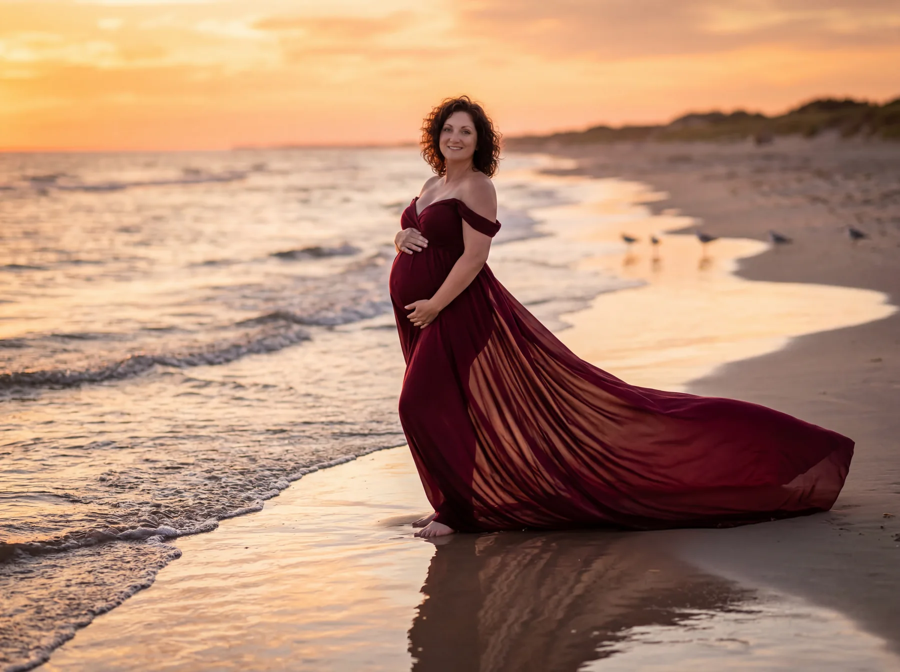 An expectant mother in a flowing burgundy gown walking beside the sea