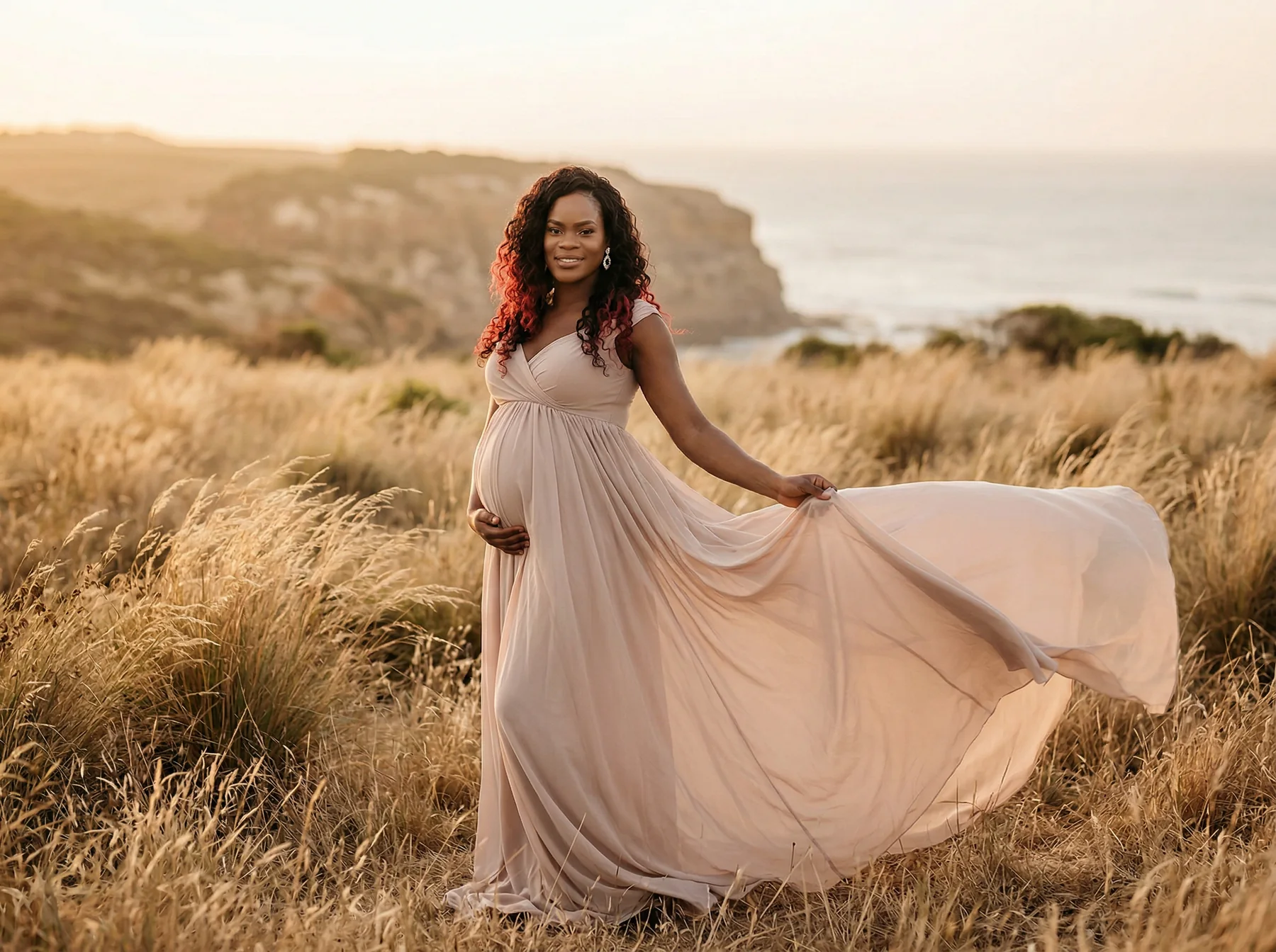 An expectant mother holding a flowing dress among coastal grasses