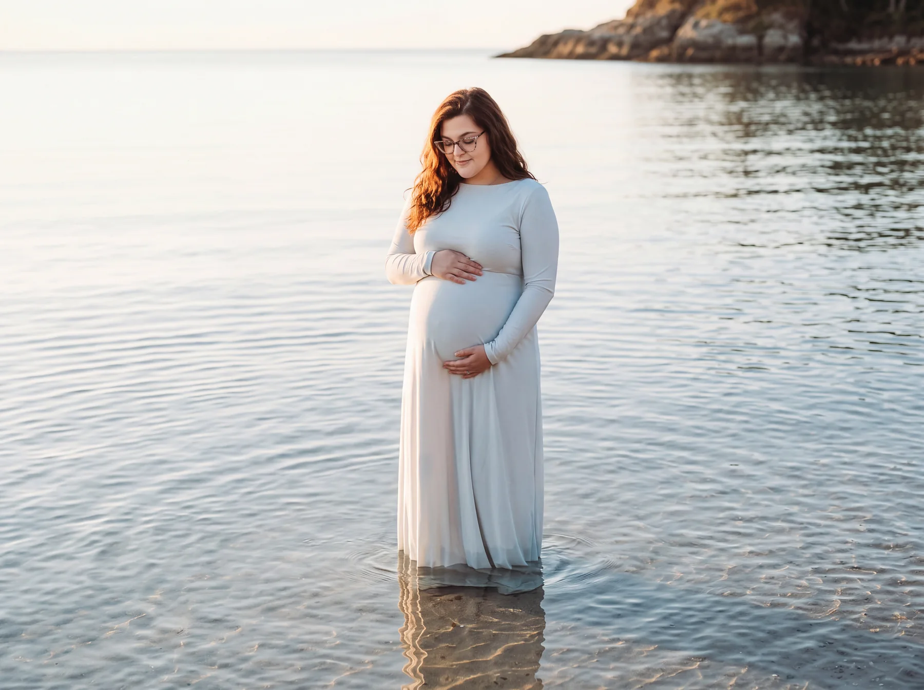 An expectant mother in a pale blue gown standing beside calm water
