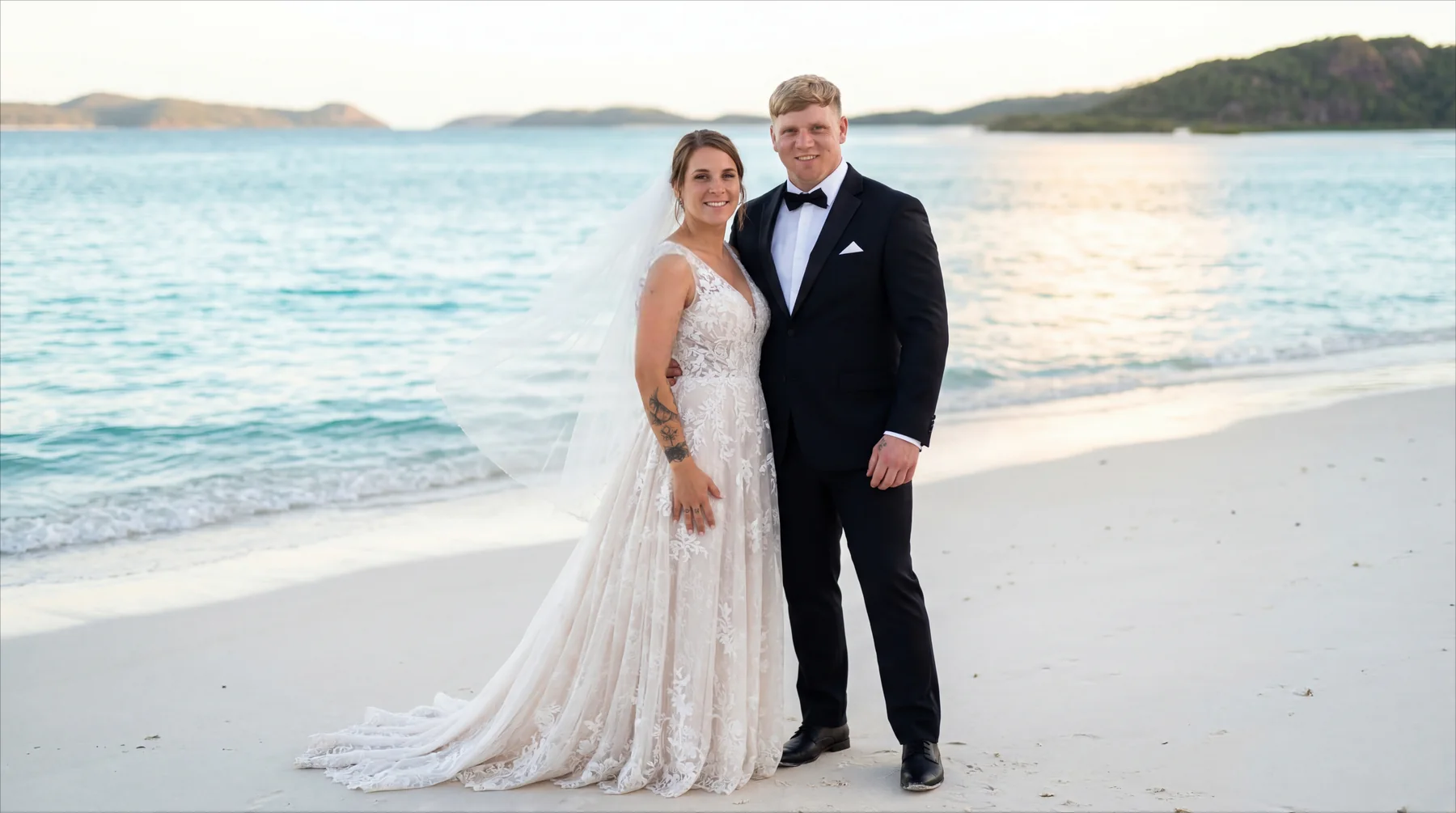 A bride and groom standing together on a bright coastal beach